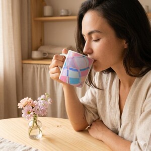 Pode incluir: Uma caneca de cerâmica branca com um padrão geométrico colorido de quadrados rosa, azuis e roxos. A caneca está sendo segurada por uma pessoa. Um pequeno vaso de vidro com flores rosa e roxas está em uma mesa de madeira.
