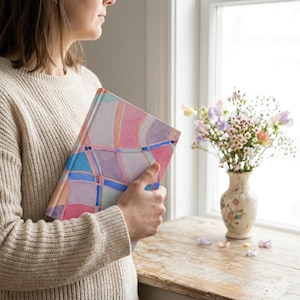 May include: A hardcover journal with a colorful abstract design in pink, blue, and purple hues. The journal is held by a person wearing a cream-colored sweater. A vase of flowers sits on a wooden surface near a window.