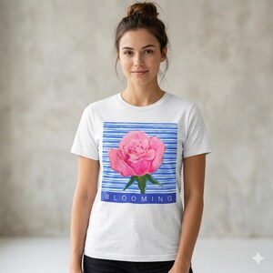 Pode incluir: Camiseta branca com uma ilustração de rosa rosa sobre um fundo listrado azul. A palavra "BLOOMING" é impressa em azul. A camiseta tem gola redonda e é de cor clara. O design apresenta uma rosa rosa com folhas verdes.