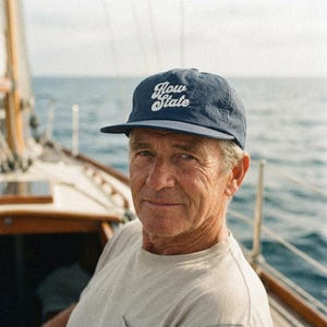 May include: A navy blue baseball cap with the text "Flow State" in white script. The cap is worn by a person on a boat, with the ocean visible in the background. The cap has a curved brim.