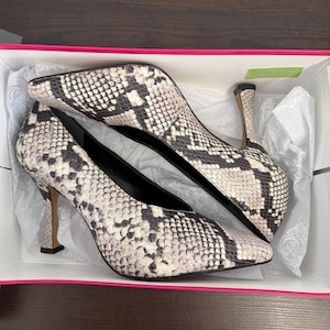 May include: A pair of high-heeled pumps with a snakeskin pattern in a box. The shoes have pointed toes and stiletto heels. The color scheme is black, white, and gray.