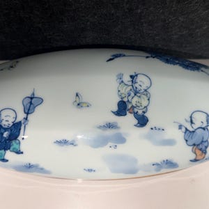 May include: A white porcelain rectangular box lid featuring blue hand-painted illustrations of three children playing outdoors. The design includes small butterflies, patches of grass, and floral elements in shades of blue and green.