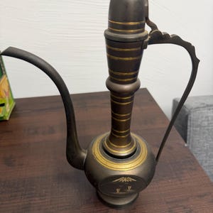 May include: An antique brass water pitcher with a long, curved spout and a handle. The pitcher has a bulbous base and a tall, cylindrical neck. Gold bands encircle the body and neck. The base features an etched design of a mountain scene.