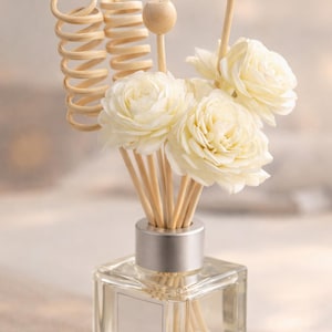 Puede incluir: Un frasco difusor de vidrio transparente con tapa plateada, lleno de varillas de madera y elementos decorativos. Las varillas sostienen varios difusores blancos con forma de flor. El fondo es de un color suave y neutro.