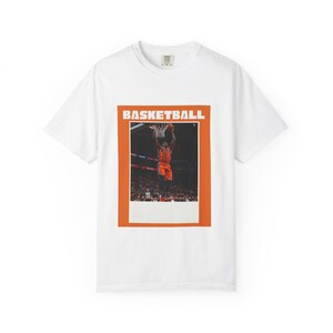 May include: White t-shirt featuring a basketball graphic. The graphic is an orange rectangle with the word "BASKETBALL" at the top. Inside the rectangle is a photo of a basketball player dunking the ball.