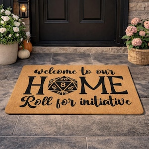 May include: A rectangular doormat with the words "welcome to our HOME Roll for initiative" in black lettering. The word "HOME" features a D20 die graphic. The mat is a natural tan color and is placed in front of a black door.