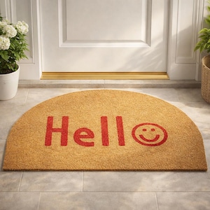 Könnte beinhalten: Eine halbkreisförmige Fußmatte aus Naturfasern. Die Matte ist beige mit dem Wort "Hello" in roten Buchstaben und einem roten Smiley. Die Matte liegt vor einer weißen Tür.