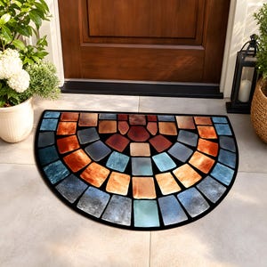 May include: A semi-circular doormat with a mosaic design. The mat features a black border and a pattern of colorful, square-shaped tiles in shades of blue, orange, brown, and gray. The doormat is placed in front of a wooden door.