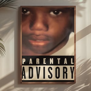 May include: A framed poster featuring a close-up of a person's face above a "Parental Advisory" label. The poster has a brown frame and is hanging on a wall. The image is lit by sunlight, with a potted plant in the foreground.