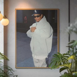 May include: Framed portrait of a person wearing a light gray hoodie, sunglasses, and a baseball cap with a white logo. The portrait is displayed on a gray wall, surrounded by green plants and hanging light fixtures.