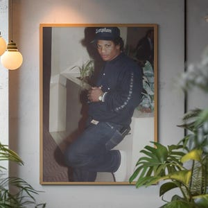 May include: A framed photograph featuring a person wearing a black jacket and a black cap with the word "Compton" on it. The person is wearing a watch and is leaning against a white surface. The photograph is in a gold frame and is hanging on a gray wall.