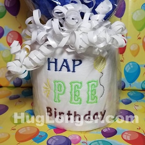 May include: A white toilet paper roll with the text "HAP PEE Birthday" embroidered on it. The roll is wrapped in clear plastic and white tissue paper.