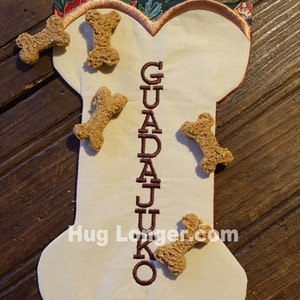 May include: A white bone-shaped stocking with the name "GUADALUPE" embroidered in brown thread. The stocking is decorated with several dog biscuits.