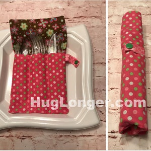 Puede incluir: Un porta cubiertos de tela de lunares rosa y verde con una solapa de tela floral marrón. El porta cubiertos está enrollado y asegurado con un botón verde. El texto "HugLonger.com" es visible en la tela.