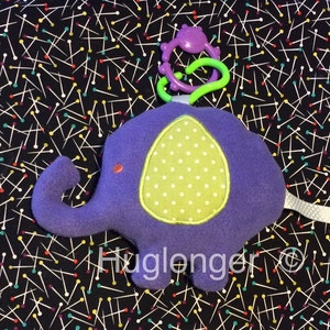 May include: Purple plush elephant toy with a green and white polka dot patch on its belly. The elephant has a colorful, plastic ring attached to its head.