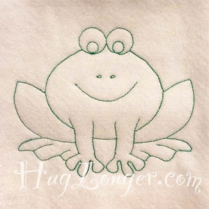 May include: A smiling frog design embroidered with green thread on a cream-colored fabric. The frog has large eyes, a wide smile, and outstretched legs. The text "HugLonger.com" is at the bottom.
