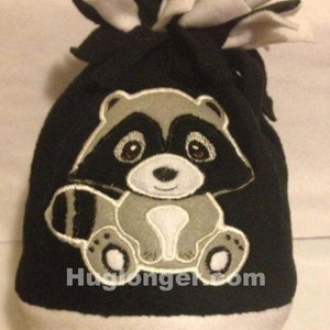 May include: Black and white fleece hat with a grey raccoon applique. The raccoon is sitting with its paws together and has a white belly and black eyes.