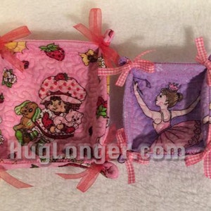 May include: Two fabric baskets with pink and purple fabric and pink and white ribbon ties. The larger basket has a strawberry print and the smaller basket has a ballerina print. The text "Huglonger.com" is visible on the baskets.