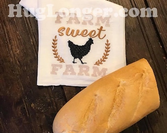 May include: A white kitchen towel with the text "Farm Sweet Farm" embroidered in brown thread. A black silhouette of a chicken is in the center of the text. The towel is lying on a wooden surface next to a loaf of white bread.