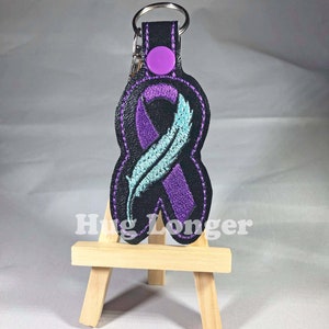 May include: Purple and teal ribbon keychain with a feather design, attached to a black leather key ring with a purple button.