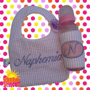 May include: Pink and white striped bib with the name Naphemia embroidered in purple. A pink and white striped bottle holder with a pink oval with the letter N embroidered in purple is attached to the bib.
