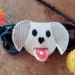 May include: A beige and brown dog-shaped cable organizer with a pink tongue. The text "NOT A PHYSICAL ITEM NOT A PHYSICAL ITEM DIGITAL FILE" is on a gold circle.
