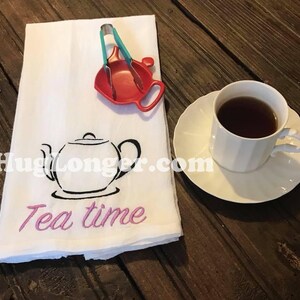 May include: White tea towel with black embroidered teapot and the words "Tea time" in pink. A red tea infuser and a white teacup with saucer are on a wooden table.
