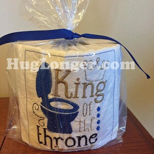 May include: A white toilet paper roll with blue and gold embroidery that reads "King of the throne". The roll is wrapped in clear plastic and tied with a blue ribbon.