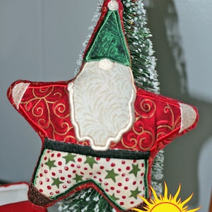 Puede incluir: Un adorno navideño de estrella roja y dorada con barba blanca y sombrero verde. El adorno está decorado con lunares rojos y blancos y estrellas verdes. El adorno está sentado en un pequeño árbol de Navidad artificial.