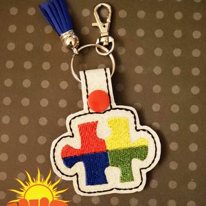 May include: A white felt keychain with a red, yellow, blue, and green puzzle piece design. The keychain has a silver key ring and a blue tassel.