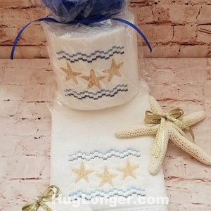 May include: A white towel with a blue and white wave pattern and three gold starfish embroidered on it. A gold starfish ornament is on the towel. A white seashell with gold glitter is on the towel. The towel is wrapped around a roll of toilet paper with a blue and white wave pattern and three gold starfish embroidered on it. The toilet paper is wrapped in blue tissue paper. A gold sticker with the text "DIGITAL FILE" is in the upper right corner of the image.