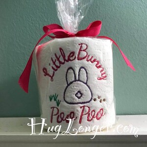 May include: A white toilet paper roll with a pink ribbon and clear plastic wrap. The roll is decorated with an embroidered bunny with the text "Little Bunny Poo Poo" and green grass.