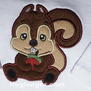 May include: A brown and tan embroidered squirrel holding a red apple. The squirrel is sitting on a white background.