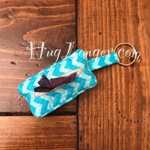 Könnte beinhalten: Ein türkisfarbener und weißer Stoff-Taschentuchhalter mit Chevron-Muster und einem silbernen Schlüsselring. Der Taschentuchhalter ist mit einem türkisfarbenen Stoffband am Schlüsselring befestigt. Der Text "Hug Longer.com" ist im Hintergrund sichtbar.