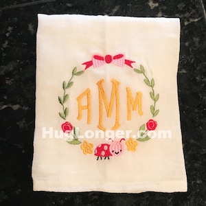 Puede incluir: Una toalla de té blanca con una corona bordada en amarillo, verde y rojo, con las letras "AMM" en el centro. Un lazo rosa con detalles rojos está en la parte superior de la corona. Una mariquita roja con manchas negras y una cara rosa está debajo de la corona.