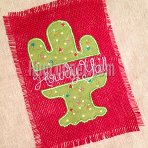 Könnte beinhalten: Ein rotes Burlap-Quadrat mit einem grünen Kaktus-Design und dem Text "Howdy Y'all", der in Weiß gestickt ist.