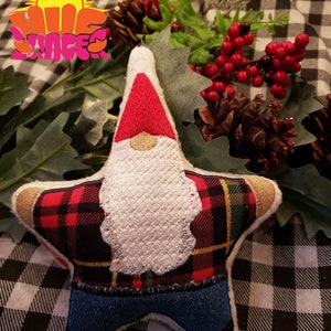 May include: A red and black plaid star-shaped gnome ornament with a white beard and blue denim pants. The ornament is sitting on a black and white checkered fabric.