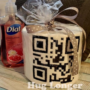 May include: A white toilet paper roll with a black and white QR code printed on it. The roll is wrapped in clear plastic with a brown ribbon and white polka dots. The text "Hug Longer" is printed below the roll.