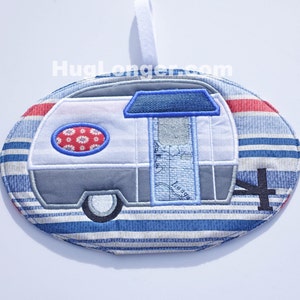May include: A blue and white striped fabric ornament with an embroidered camper van design. The camper van is gray with a blue roof and a blue door. The ornament has a loop for hanging.