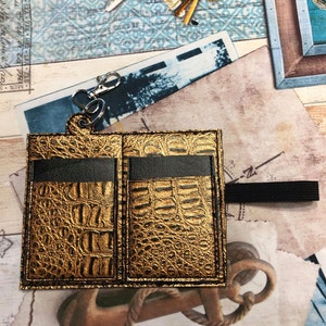 May include: A gold-colored leather card holder with a crocodile pattern. The holder has two slots for cards and a black elastic strap.