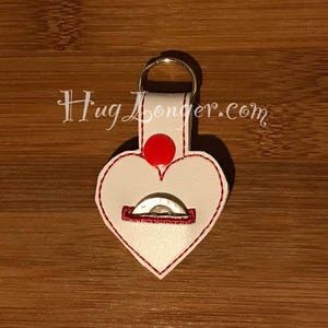 May include: White leather heart-shaped keychain with a red button and a red stitched pocket for a coin. The keychain has a gold metal ring and the text "HugLonger.com" is printed on the top.