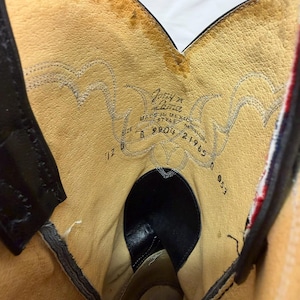 K&ouml;nnte beinhalten: Das Innere eines Cowboystiefels aus Leder mit beigefarbenem Futter und Ziern&auml;hten. Der aufgedruckte Text enth&auml;lt Tony Lama, Made in Mexico, die Modellnummer 21965 und die Gr&ouml;&szlig;e 7 1/2 B. Der Stiefel hat schwarze Lederakzente und einen rot-wei&szlig;en Rand.
