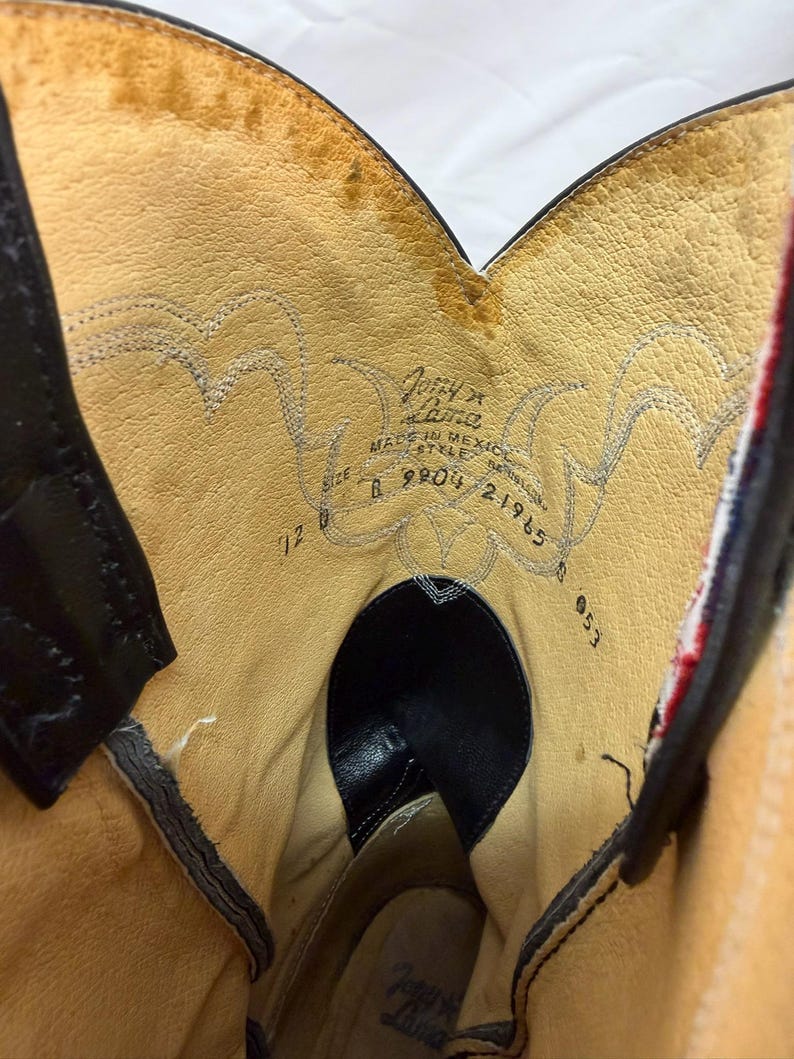 K&ouml;nnte beinhalten: Das Innere eines Cowboystiefels aus Leder mit beigefarbenem Futter und Ziern&auml;hten. Der aufgedruckte Text enth&auml;lt Tony Lama, Made in Mexico, die Modellnummer 21965 und die Gr&ouml;&szlig;e 7 1/2 B. Der Stiefel hat schwarze Lederakzente und einen rot-wei&szlig;en Rand.