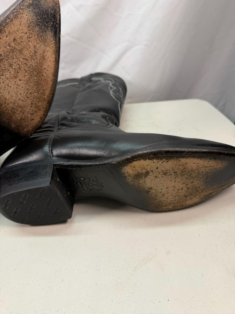 K&ouml;nnte beinhalten: Ein Paar schwarze Cowboystiefel aus Leder mit spitzer Zehenpartie, Blockabsatz und dekorativen wei&szlig;en Ziern&auml;hten am Schaft. Die Sohlen weisen Gebrauchsspuren mit strukturierter Oberfl&auml;che auf und der Absatz zeigt ein wiederkehrendes Logomuster.