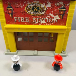 May include: A vintage Fisher-Price Play Family Fire Station toy. The building is yellow and brown with a red brick facade and the words "FIRE STATION" printed on the front. Two small firefighter figures are in front of the building.