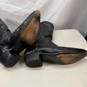 K&ouml;nnte beinhalten: Ein Paar schwarze Leder-Cowboystiefel mit spitzer Zehenpartie, Blockabs&auml;tzen und hellen Ziern&auml;hten am Schaft. Die Sohlen weisen Gebrauchsspuren auf und die Abs&auml;tze sind an der Unterseite mit einem strukturierten Muster versehen.