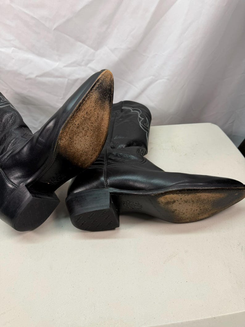 K&ouml;nnte beinhalten: Ein Paar schwarze Leder-Cowboystiefel mit spitzer Zehenpartie, Blockabs&auml;tzen und hellen Ziern&auml;hten am Schaft. Die Sohlen weisen Gebrauchsspuren auf und die Abs&auml;tze sind an der Unterseite mit einem strukturierten Muster versehen.