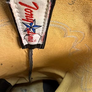 K&ouml;nnte beinhalten: Das Innere eines schwarzen Leder-Cowboystiefels mit beigem Futter. Ein wei&szlig;es Stoffetikett ist oben aufgen&auml;ht und zeigt den roten Schriftzug Tony Lama sowie ein blaues Sternsymbol. Die Zahl 3 ist in das beige Leder nahe der Oberkante eingepr&auml;gt.