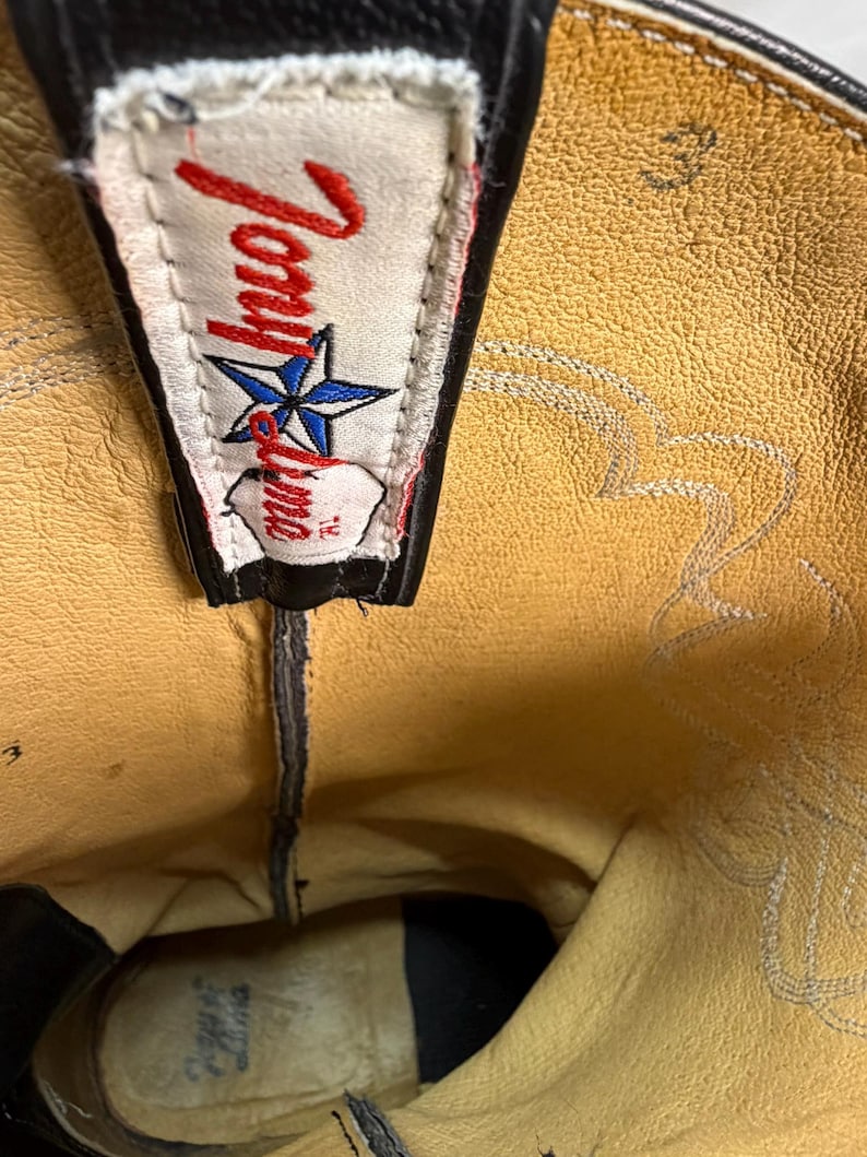K&ouml;nnte beinhalten: Das Innere eines schwarzen Leder-Cowboystiefels mit beigem Futter. Ein wei&szlig;es Stoffetikett ist oben aufgen&auml;ht und zeigt den roten Schriftzug Tony Lama sowie ein blaues Sternsymbol. Die Zahl 3 ist in das beige Leder nahe der Oberkante eingepr&auml;gt.