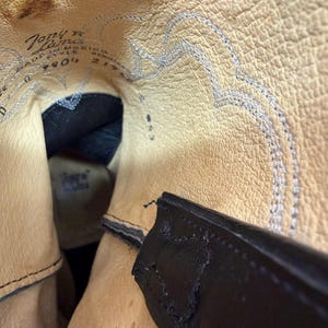 K&ouml;nnte beinhalten: Das Innere eines Stiefels mit beigefarbenem Lederfutter und strukturierter Oberfl&auml;che. Der eingepr&auml;gte Text lautet Tony Lama, Made in Mexico, Style 9804 und 21768. Eine schwarze Lederlasche ist sichtbar, sowie silberne Ziern&auml;hte am Schaft.